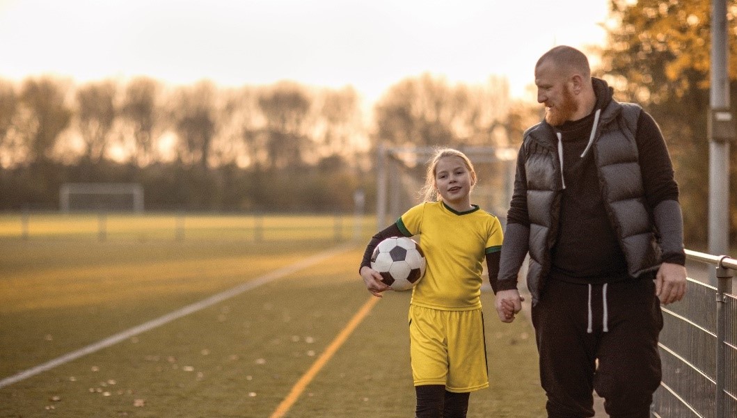 A young soccer player in a yellow uniform holds a ball and walks hand-in-hand with an adult on a tree-lined field.