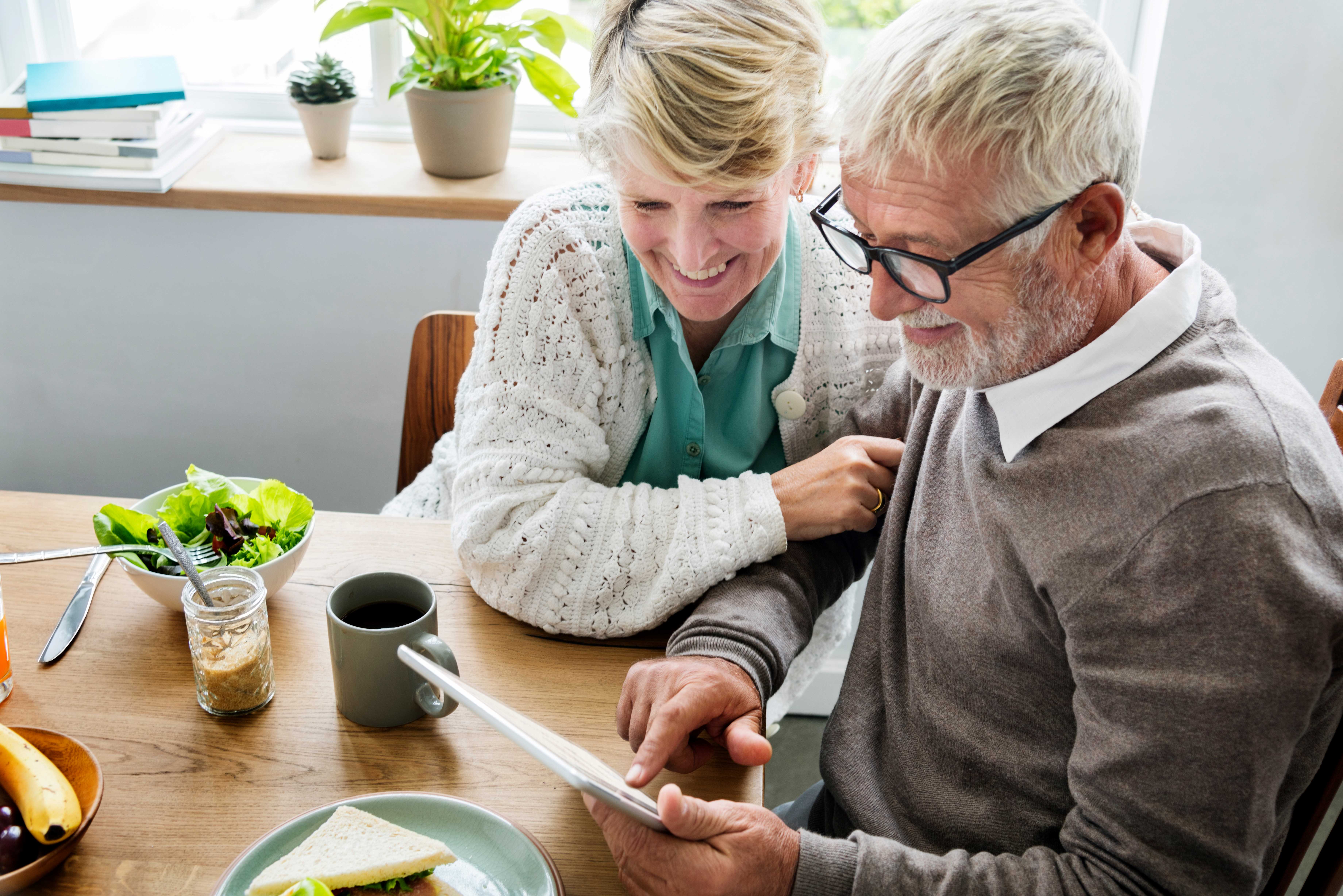 Photo of man and woman at table with healthy food options on it, they're looking at a tablet the man is holding and smiling.