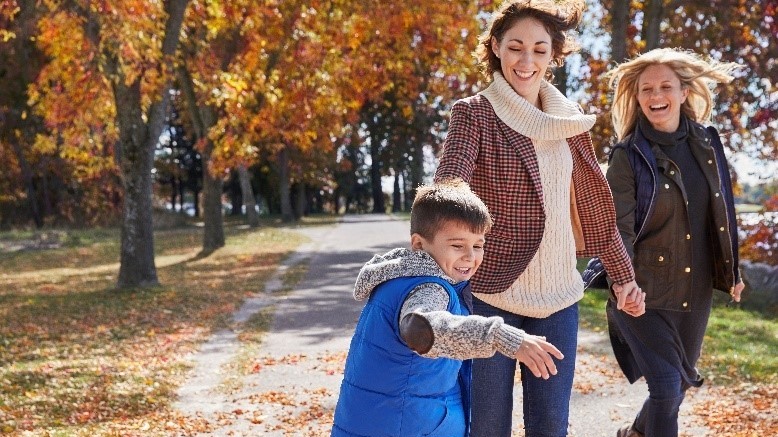 Two smiling women wearing coats are out for a neighborhood walk with a boy. Orange leaves from the trees lining the sidewalk cover the path..