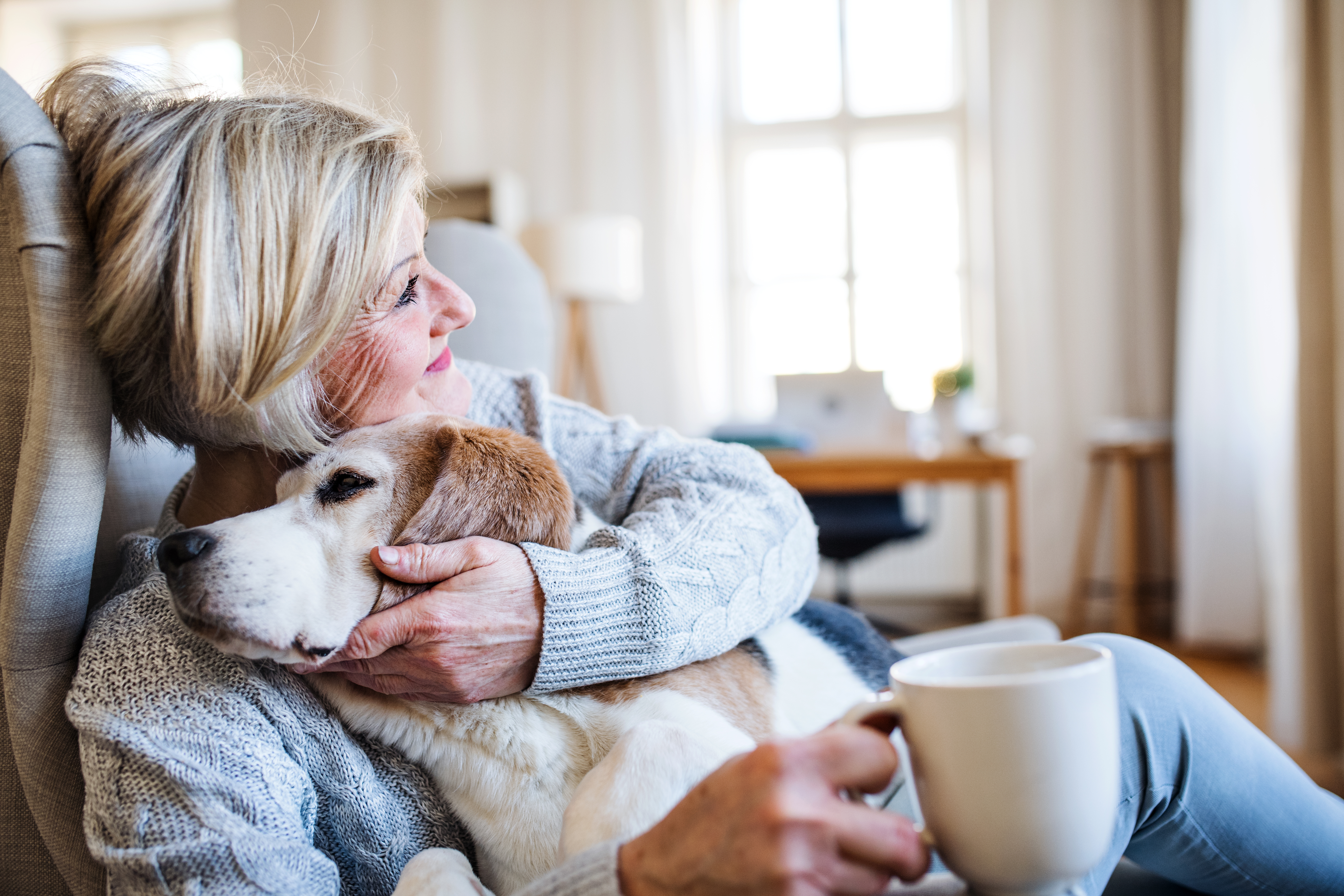 Smiling woman with white hair sits on the floor in her home with coffee cup in one hand as she hugs her dog as it snuggles into her chest..
