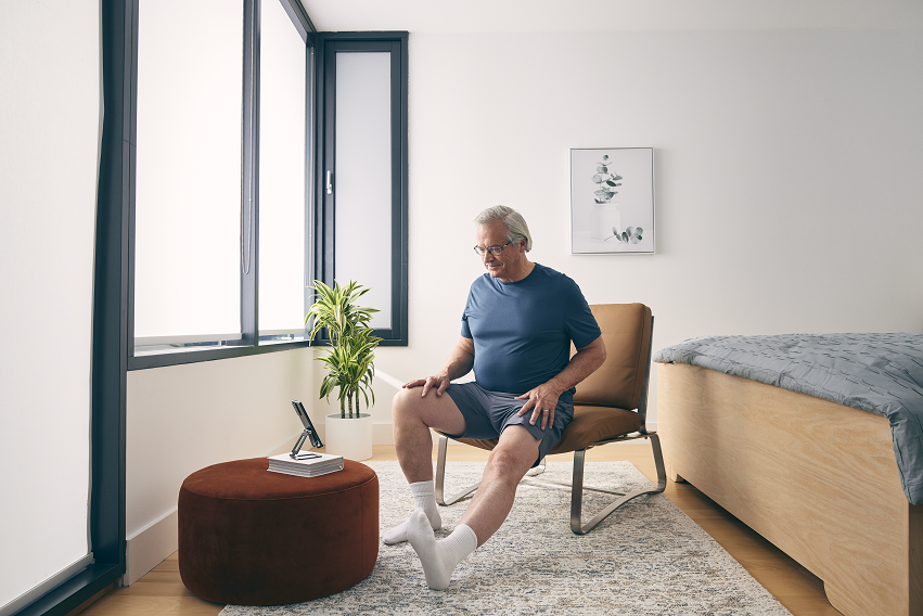 Man sitting in chair with phone propped up so he can follow along with a leg extention excercise 