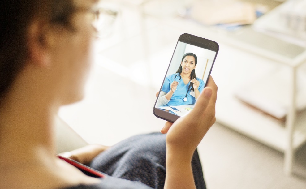 A person holds a smartphone while seated indoors; on the phone screen, a healthcare professional wearing scrubs appears during a video call, with papers and a pen visible on a desk.