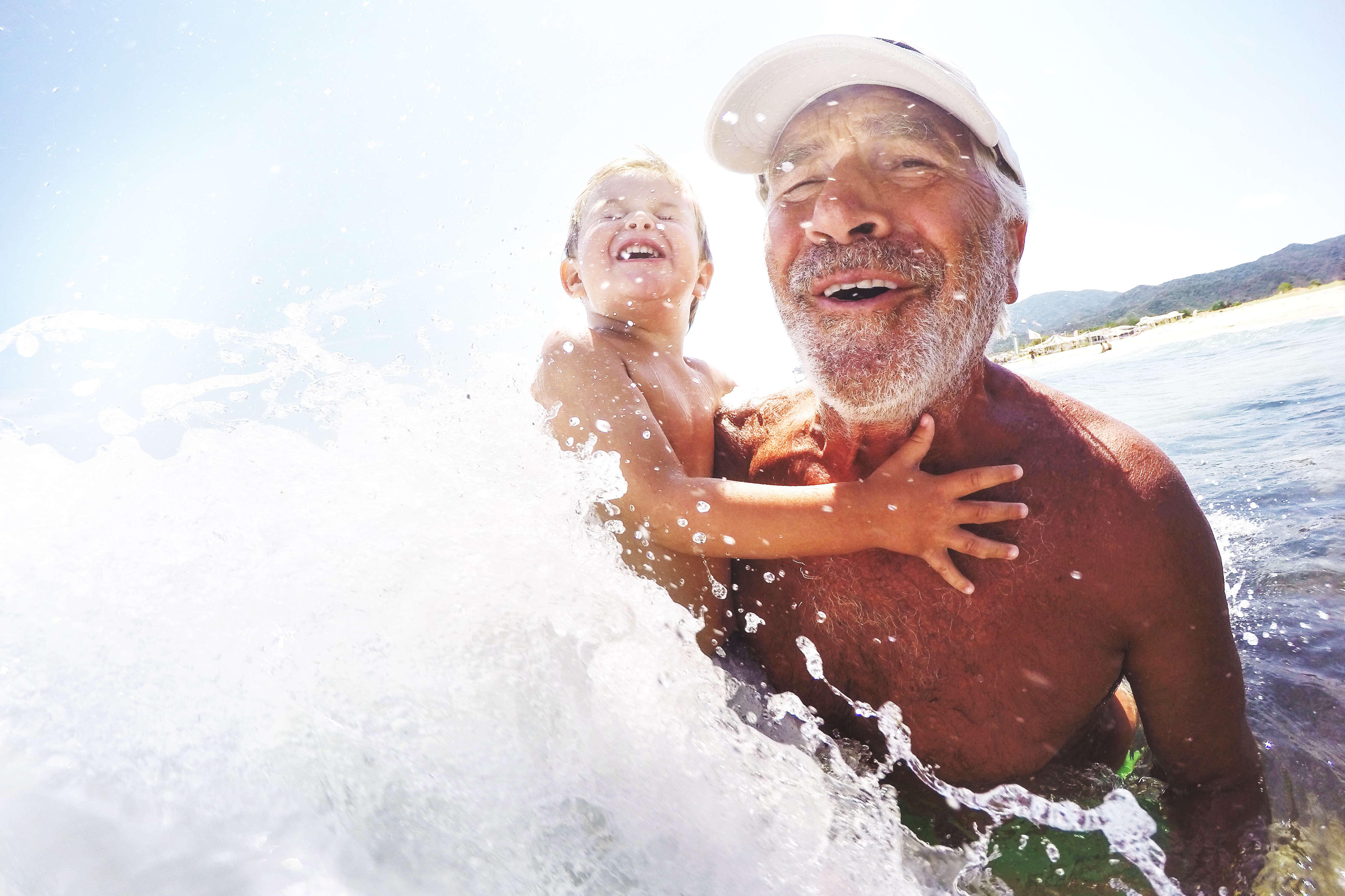 A wave splashes against a smiling grandfather carrying his happy grandchild into the water at a beach