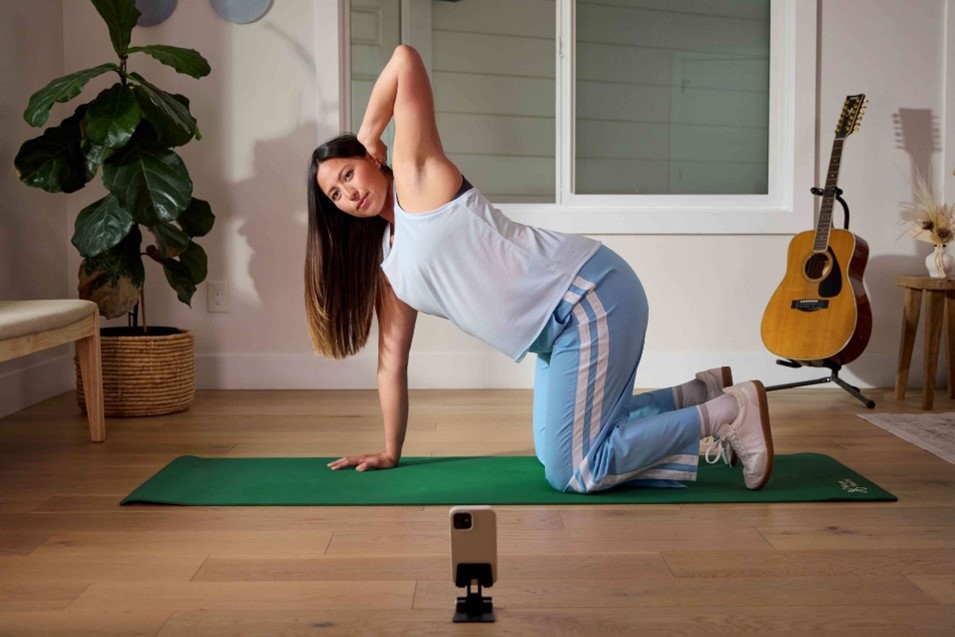 Woman does stretch on a yoga mat with her phone proped up facing her.