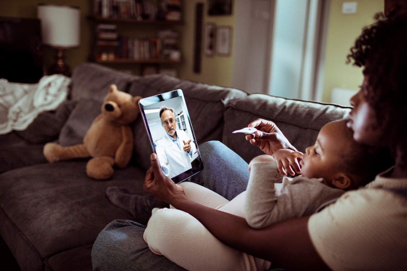 Serious woman with a small child in her lap holds a tablet in front of them as they video call with a doctor .