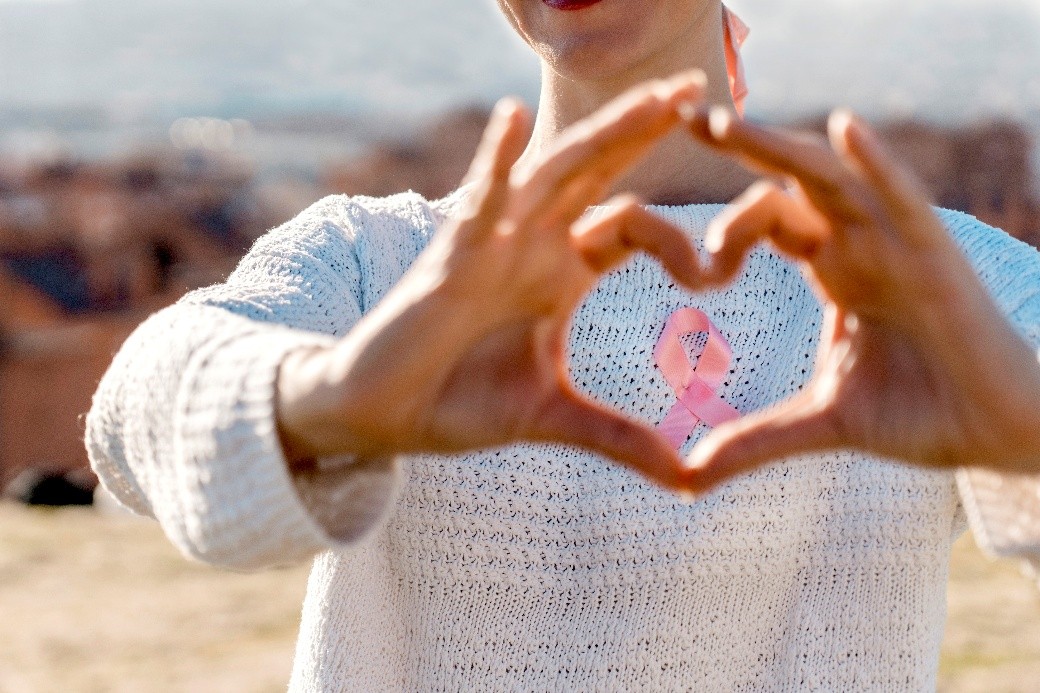 Person wearing a pink breast cancer awareness ribbon and forming a heart shape with both hands.