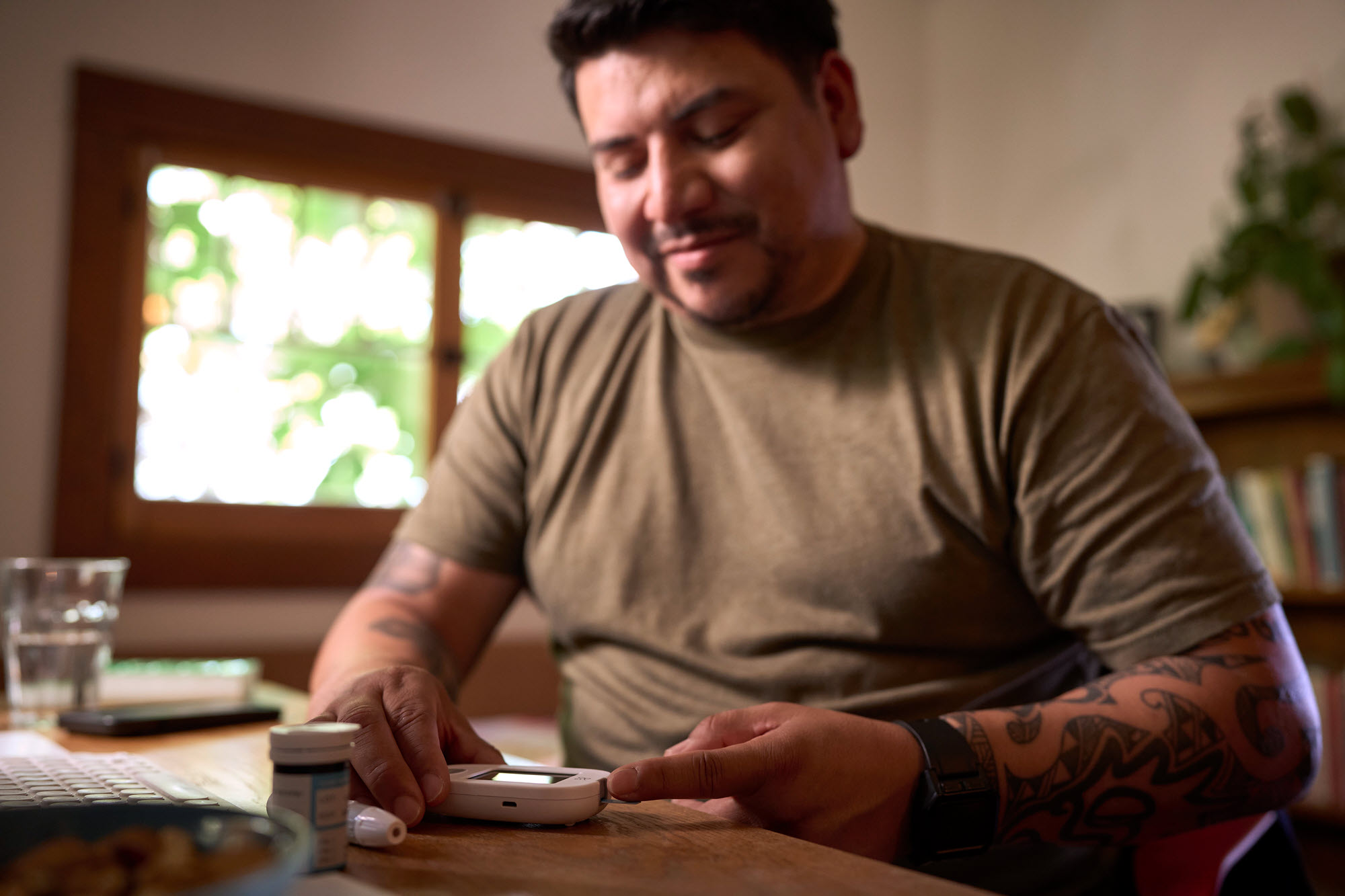 A man sits at a table in a warmly lit room, using a blood glucose monitor,. He has tattoos on his arms and wears a smartwatch and a khaki t-shirt. Sunlight streams in through a window behind him, with greenery visible outside.